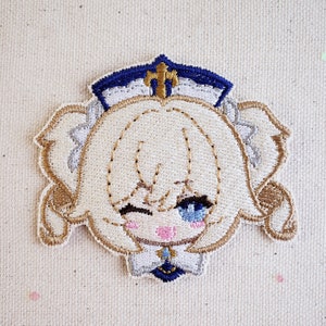 May include: Embroidered patch of a cartoon character with blonde hair, blue eyes, and a pink blush. The character is wearing a white and blue hat with a gold cross on top.