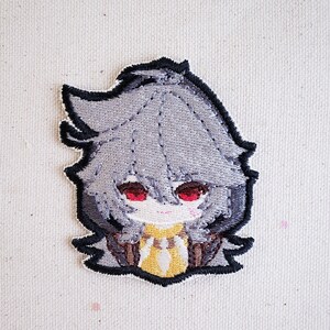 May include: Embroidered patch featuring a cartoon character with gray hair, red eyes, and a yellow and brown outfit.