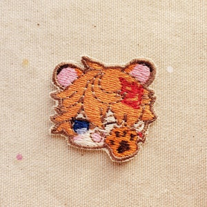 May include: Embroidered patch featuring a cartoon tiger cub with orange fur, blue eyes, and a paw print. The patch has a brown border and is stitched on a beige fabric background. The tiger cub has a red bandage on its head.