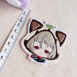 May include: Embroidered iron-on patch featuring a cartoon character with brown cat ears, a green leaf, and a white background.