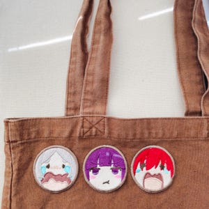 Puede incluir: Un bolso de tela marrón con tres parches bordados circulares. El parche de la izquierda muestra un personaje llorando con pelo blanco, el central muestra un personaje con pelo morado y expresión neutra, y el de la derecha muestra un personaje llorando con pelo rojo.