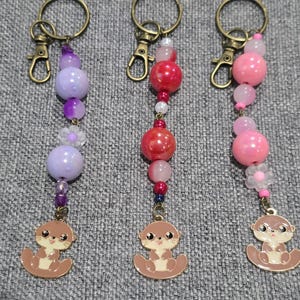 May include: Three beaded keychains with otter charms. One keychain features purple beads, another red, and the third pink. Each keychain has a metal clasp and a small otter charm at the bottom.