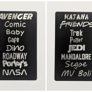 May include: Two black rectangular signs with silver lettering. The left sign has the words "AVENGER", "Comic", "Baby", "Cafe", "Dino", "ROADWAY", "Porky's", and "NASA". The right sign has the words "KATANA", "FRIENDS", "Trek", "Potter", "JEDI", "MANDALORE", "Segoe", "MV Boli".