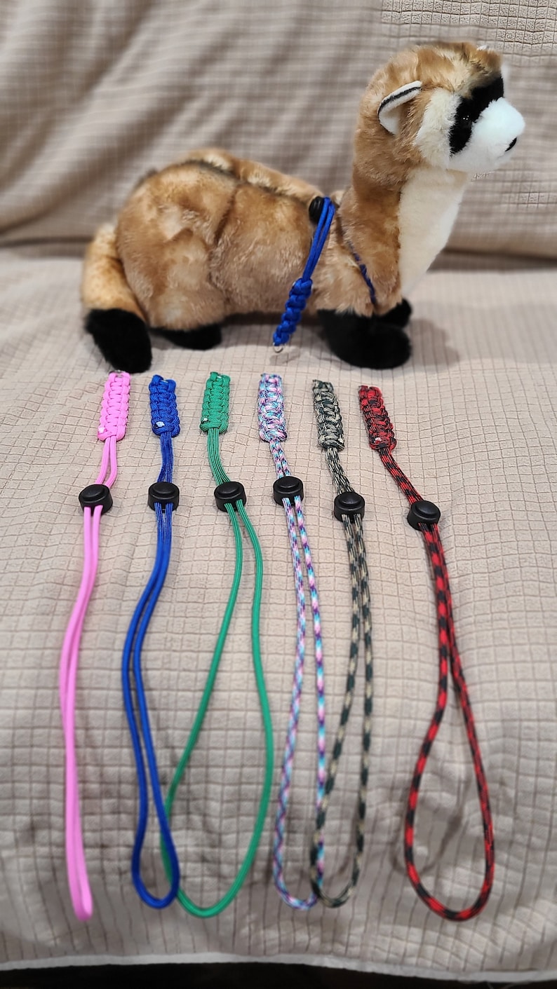 May include: A plush ferret toy with a blue harness is displayed with six colorful paracord leashes. The leashes are pink, blue, green, multicolored, gray, and red. Each leash has a black adjustable slider.