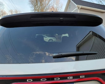 May include: A white Dodge SUV with a black rear spoiler and a rear window wiper. The rear window is tinted and reflects the sky and a house.