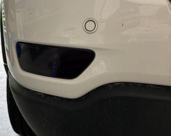 May include: White car with a black rear bumper and a single exhaust pipe. The car has a black rear reflector and a small, round sensor on the bumper.