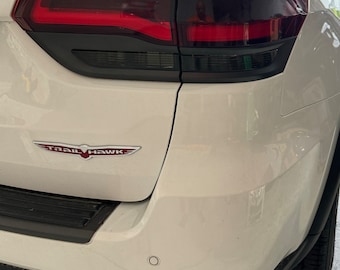 May include: White Jeep Trailhawk with a red and silver Trailhawk emblem on the rear. The rear lights are tinted black.