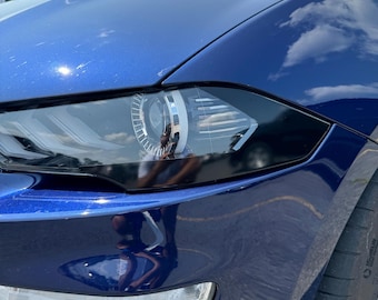 May include: A close-up of a blue car's front headlight. The headlight is on and the car is parked on a paved surface.