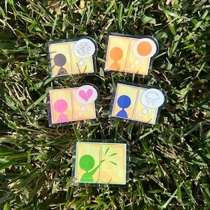 May include: Five colorful acrylic pins with a person silhouette and thought bubbles. The thought bubbles contain a heart, a sun, a rain cloud, a tangled line, and a burst of light. The pins are on a bed of green grass.