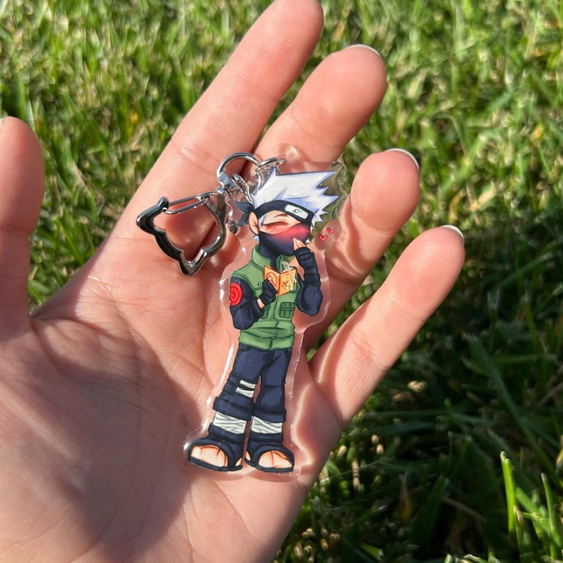 K&ouml;nnte beinhalten: Ein klarer Acryl-Schl&uuml;sselanh&auml;nger mit einer Cartoonfigur mit wei&szlig;en Haaren, einer schwarzen Maske und einer gr&uuml;nen Weste. Die Figur h&auml;lt ein Buch und hat ein rotes Symbol am linken Arm. Der Schl&uuml;sselanh&auml;nger hat einen silbernen Verschluss.