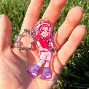 K&ouml;nnte beinhalten: Ein klarer Acryl-Schl&uuml;sselanh&auml;nger mit einem pinkhaarigen Anime-Charakter in einem roten Oberteil und Shorts, der eine kleine Waffe h&auml;lt. Der Schl&uuml;sselanh&auml;nger hat einen silbernen bl&uuml;tenf&ouml;rmigen Anh&auml;nger.