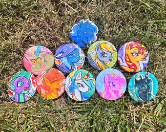 May include: A collection of ten circular pin-back buttons featuring colorful illustrations of various animated pony and creature characters. Each button displays a unique character with distinct facial expressions, vibrant color palettes, and artistic line work.