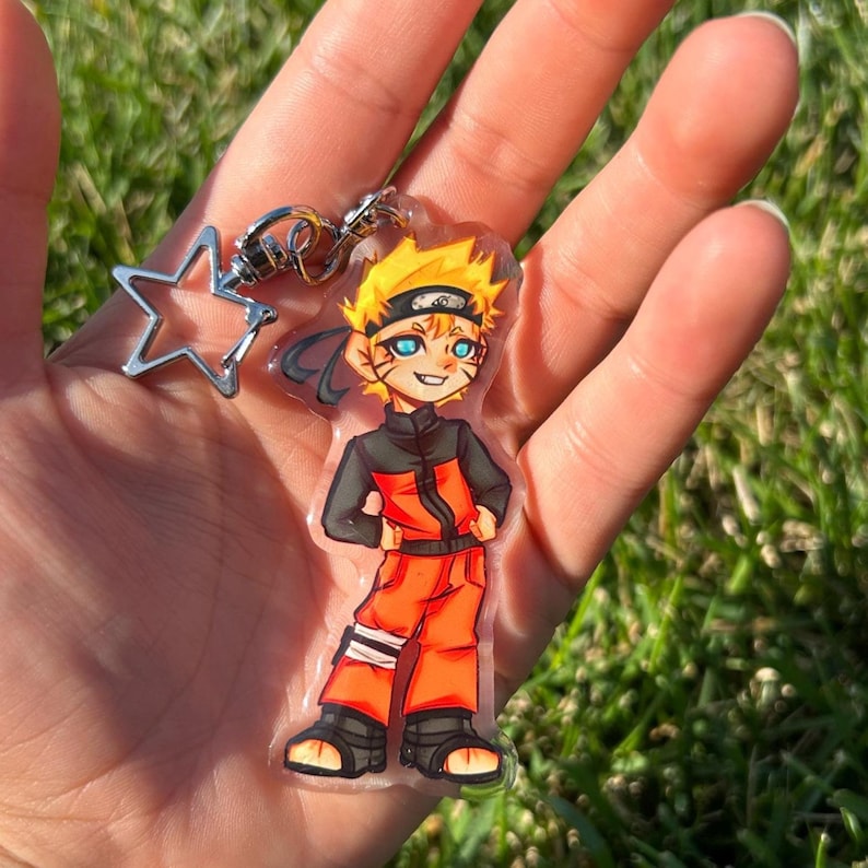 K&ouml;nnte beinhalten: Ein klarer Acryl-Schl&uuml;sselanh&auml;nger mit einer Cartoonfigur mit stacheligen gelben Haaren, blauen Augen und orangefarbener und schwarzer Kleidung. Der Schl&uuml;sselanh&auml;nger enth&auml;lt einen silbernen sternf&ouml;rmigen Anh&auml;nger und einen Metallverschluss.