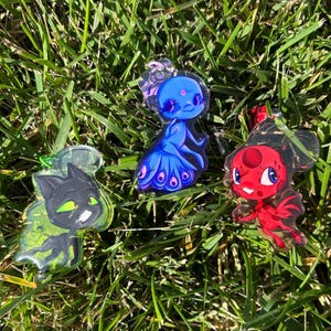 May include: Three colorful acrylic charms featuring cartoon characters. One is black with green accents, one is blue with a peacock-inspired design, and the last is red with black accents. The charms are displayed on a bed of green grass.
