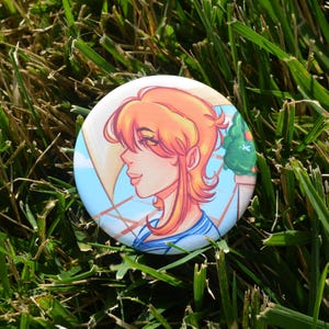 May include: A round button with an illustration of a person with orange hair and a blue and white striped shirt. The background is light blue with a sailboat and a green tree. The button is set on a bed of green grass.