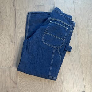 Könnte beinhalten: Eine dunkelblaue Jeanshose im Carpenter-Stil mit weißen Nähten. Die Jeans hat eine große Gesäßtasche und eine Hammerschlaufe.