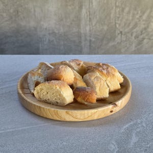 May include: A wooden plate displays several pieces of freshly baked bread. The bread has a golden-brown crust and a soft, light interior. The plate is made of light-coloured wood and has a natural grain pattern.