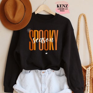 May include: Black sweatshirt with the text "SPOOKY season" in orange and yellow letters. The sweatshirt is hanging on a hanger and is styled with a brown hat and a straw bag.