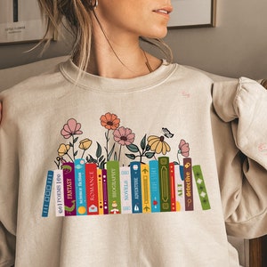 Puede incluir: Una sudadera beige con un gr&aacute;fico de una pila de libros con diferentes g&eacute;neros, incluyendo poes&iacute;a, fantas&iacute;a, ciencia ficci&oacute;n, romance, biograf&iacute;a, novelas, aventura y detectives. Los libros est&aacute;n rodeados de flores y una mariposa. El texto "Sand" est&aacute; impreso debajo del gr&aacute;fico.