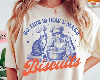 May include: A cream-colored t-shirt with a blue illustration of a cat and a woman wearing chef hats, baking biscuits. The text "So this is how u make biscuits" is printed in blue and orange.
