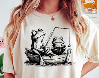 May include: A white t-shirt featuring a black and white illustration of two frogs in a boat. One frog is fishing while the other is reading a book.