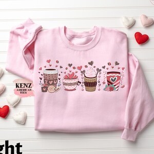 May include: Light pink sweatshirt with a graphic of five different coffee cups with hearts and the text "KENZ AMERICAN TEES".