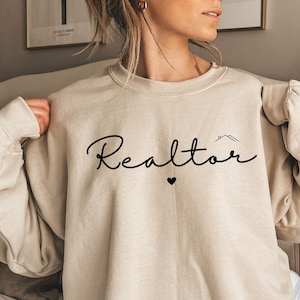May include: A beige sweatshirt with the word "Realtor" in black script with a small house icon and a heart above the word. The text "Sand" is visible at the bottom of the image.