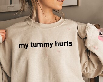 May include: A sand-colored sweatshirt with the text "my tummy hurts" printed in black. The sweatshirt is a relaxed fit and has a crew neck.