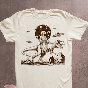 May include: A beige t-shirt with a brown graphic print of Jesus riding a dinosaur with a UFO in the background. The graphic also includes a Bigfoot figure and a moon with stars.