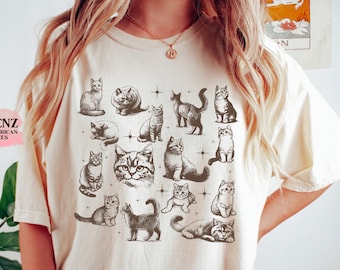 May include: A cream-colored t-shirt with a vintage-style illustration of multiple cats in various poses. The cats are all black and white and are surrounded by small stars.