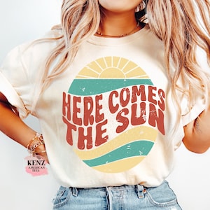 Pode incluir: Uma camiseta branca com um gr&aacute;fico retr&ocirc; de um sol com o texto "Here Comes The Sun" em letras vermelhas e amarelas.