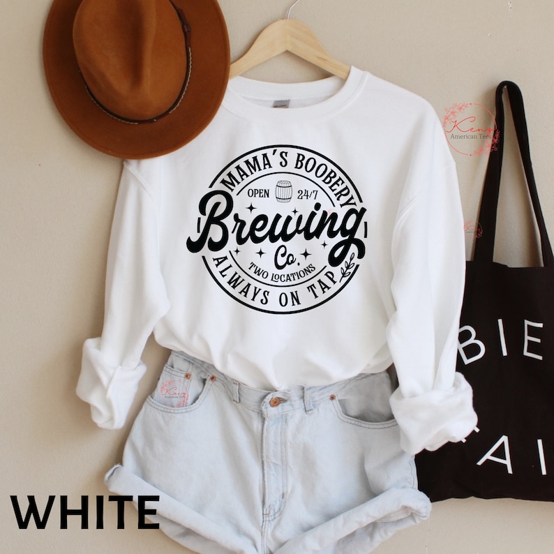 Puede incluir: Sudadera blanca con un dise&ntilde;o gr&aacute;fico en blanco y negro que dice "Mama's Boobery Brewing Co. Always on Tap" con un icono de jarra de cerveza. La sudadera se lleva sobre unos pantalones cortos de mezclilla azul claro.