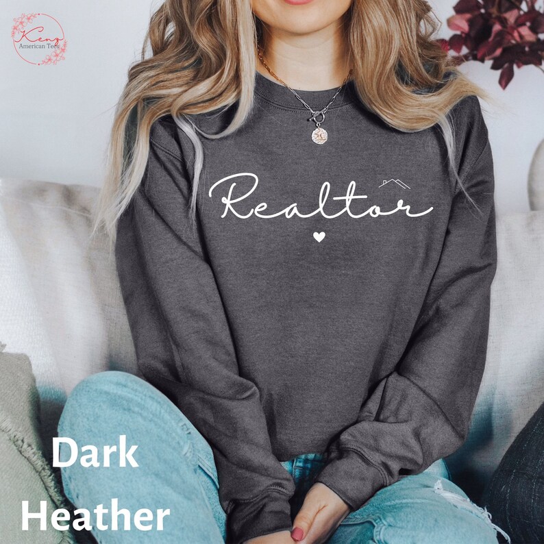 May include: A dark heather grey sweatshirt with the word "Realtor" written in white cursive with a small heart and a house icon above the word.