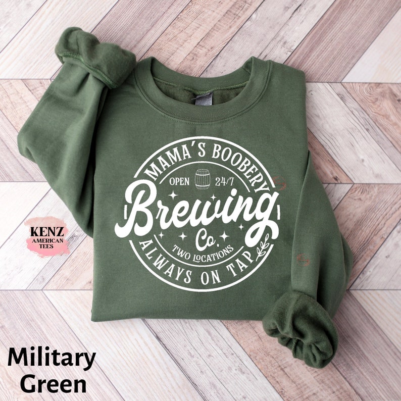 Puede incluir: Una sudadera verde con un gr&aacute;fico circular blanco que dice "Mama's Boobery Brewing Co. Always On Tap" con un barril, estrellas y las palabras "Open 24/7" y "Two Locations".