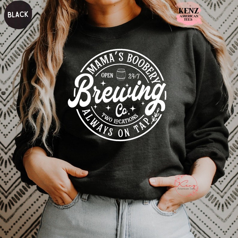 Puede incluir: Sudadera negra con un gr&aacute;fico circular blanco que dice "Mama's Boobery Brewing Co. Always on Tap" con un peque&ntilde;o icono de barril y las palabras "Open 24/7" dentro del c&iacute;rculo.