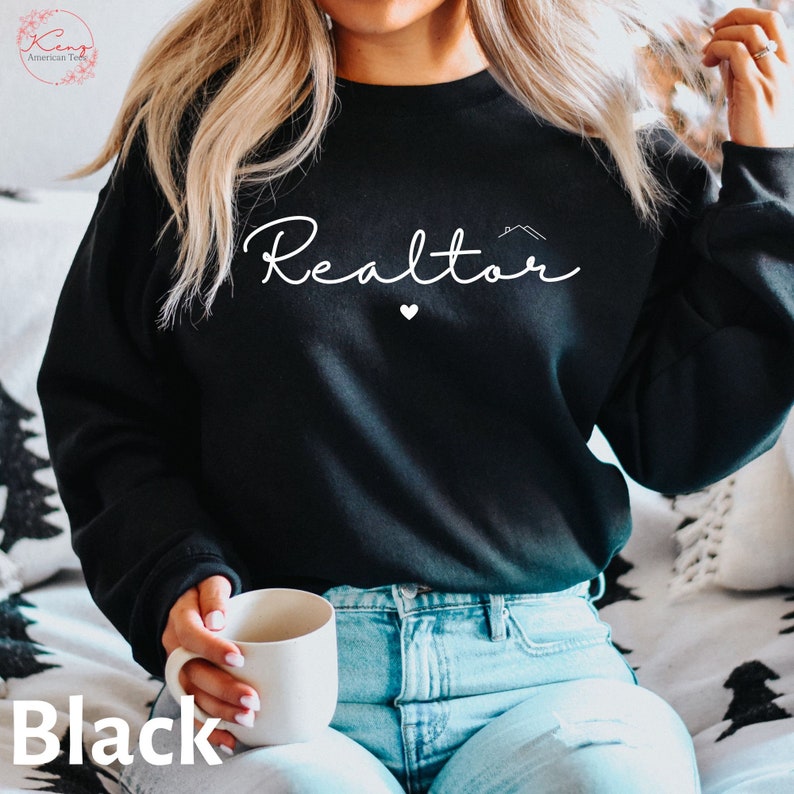 May include: Black sweatshirt with the text "Realtor" in white cursive lettering, a small house icon, and a heart. The model is wearing blue jeans.