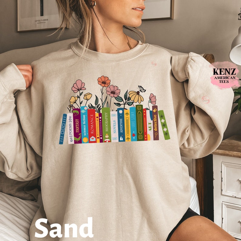 Puede incluir: Una sudadera beige con un gr&aacute;fico de una pila de libros con diferentes g&eacute;neros, incluyendo poes&iacute;a, fantas&iacute;a, ciencia ficci&oacute;n, romance, biograf&iacute;a, novelas, aventura y detectives. Los libros est&aacute;n rodeados de flores y una mariposa. El texto "Sand" est&aacute; impreso debajo del gr&aacute;fico.