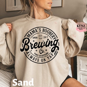 Puede incluir: Una sudadera beige con un estampado gr&aacute;fico negro que dice "Mama's Boobery Brewing Co. Always On Tap" con un icono de barril de cerveza y estrellas. El texto est&aacute; en una fuente de estilo vintage.