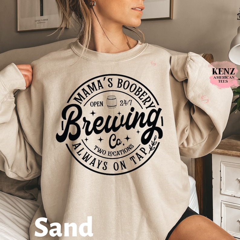 Puede incluir: Una sudadera beige con un estampado gr&aacute;fico negro que dice "Mama's Boobery Brewing Co. Always On Tap" con un icono de barril de cerveza y estrellas. El texto est&aacute; en una fuente de estilo vintage.