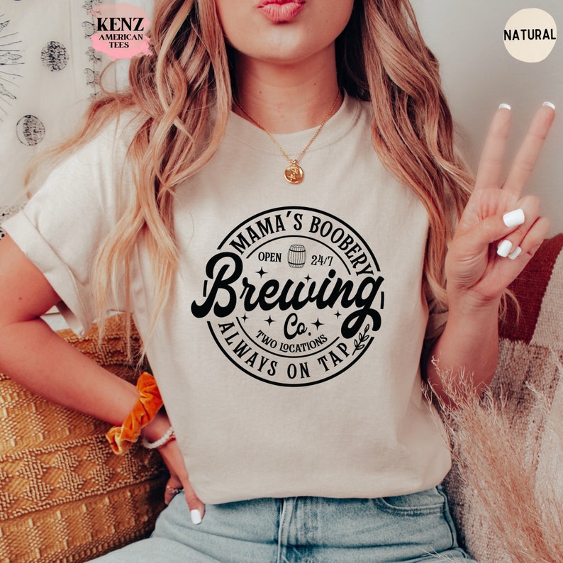 Puede incluir: Una camiseta blanca con un gr&aacute;fico negro que dice "Mama's Boobery Brewing Co.  Two Locations Always On Tap". El gr&aacute;fico es un c&iacute;rculo con un barril y estrellas.