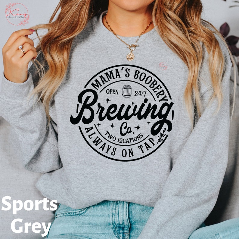 Puede incluir: Una sudadera gris con un estampado gr&aacute;fico negro que dice "Mama's Boobery Brewing Co. Always On Tap" con un icono de barril y estrellas.