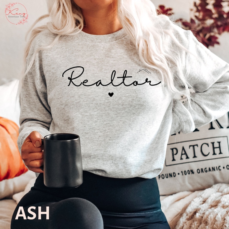 May include: A gray sweatshirt with the word "Realtor" written in black cursive with a small heart below it. The sweatshirt is being worn by a person holding a black coffee mug.