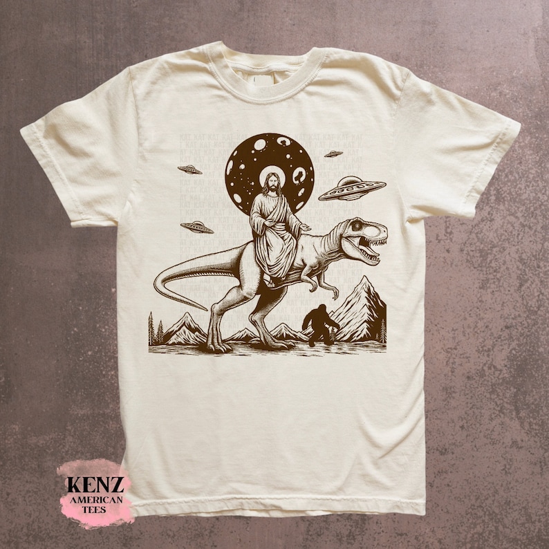 May include: A beige t-shirt with a brown graphic print of Jesus riding a dinosaur with a UFO in the background. The graphic also includes a Bigfoot figure and a moon with stars.