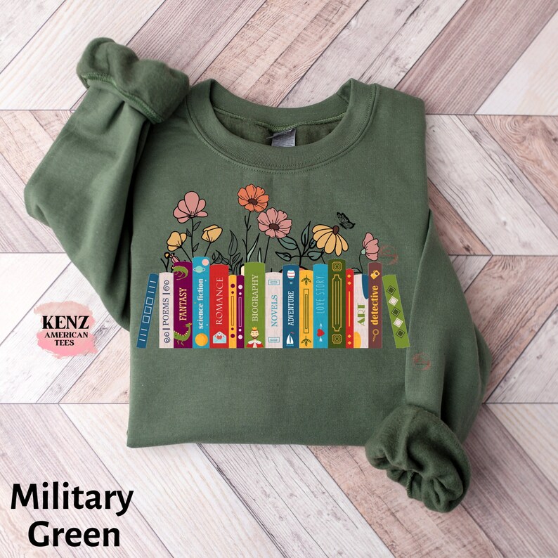 Puede incluir: Una sudadera verde con un gr&aacute;fico de una pila de libros con diferentes g&eacute;neros, incluyendo poes&iacute;a, fantas&iacute;a, ciencia ficci&oacute;n, romance, biograf&iacute;a, novelas, aventura, historia de amor, arte y detectives. Los libros est&aacute;n rodeados de flores.