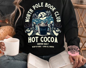 May include: Black sweatshirt with a graphic that reads "North Pole Book Club Hot Cocoa." The design includes a cup of cocoa, reindeer, and a sleigh. The text "Served Daily, Warp Up Here, Open All Winter" is also included.