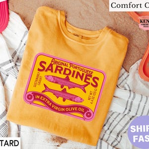 May include: Mustard-colored t-shirt with a pink and yellow graphic of a sardine can, reading "Original Portuguese Sardines." The shirt also features the text "Sustainably Caught" and "In Extra Virgin Olive Oil." The shirt is next to a pink cap, sunglasses, and orange sandals.