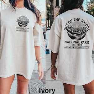 Puede incluir: Camiseta de gran tamaño de color marfil con un estampado gráfico negro de una escena de montaña dentro de un círculo con el texto "Out of the Woods National Park Est. 1989 Are we in the clear yet?"