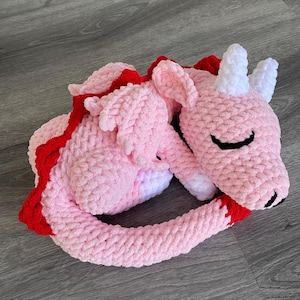 Könnte beinhalten: Ein handgefertigtes, rosa gehäkeltes Drachenplüschtier mit roten Akzenten. Der Drache ist zusammengerollt, mit weißen Hörnern und einem schwarz umrandeten Auge. Das Plüschtier ist aus weichem, strukturiertem Garn gefertigt.
