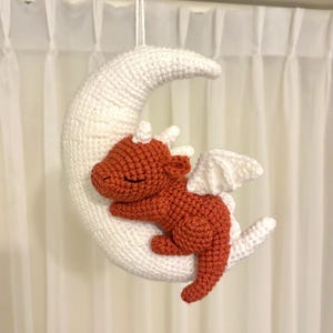 Könnte beinhalten: Gehäkeltes Baby-Mobile mit einem schlafenden orangefarbenen Drachen auf einem weißen Halbmond. Der Drache hat weiße Flügel und kleine Hörner. Das Mobile wird an einer weißen Schnur vor einem weißen Vorhang aufgehängt.