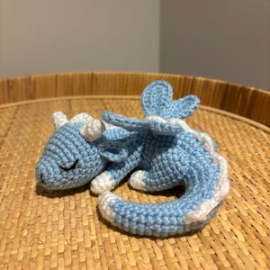 May include: A crocheted blue and white dragon figurine. The handmade dragon is lying down with its eyes closed. The dragon has white horns, white accents, and a white belly. The dragon is resting on a woven, light brown surface.
